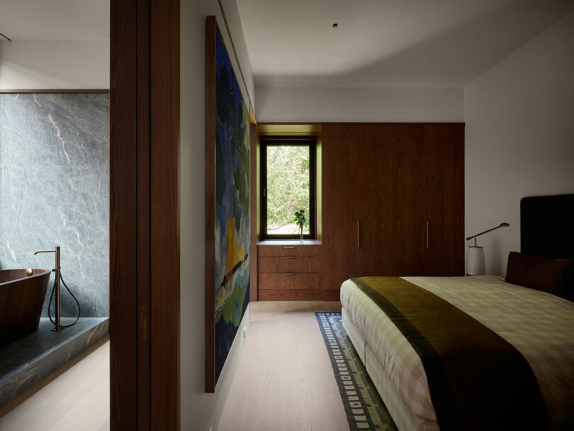 Modern bedroom designed by Atelier Echelle features a large bed, wooden wardrobe, abstract painting on the wall, and a view of an adjacent bathroom with a stone bathtub.