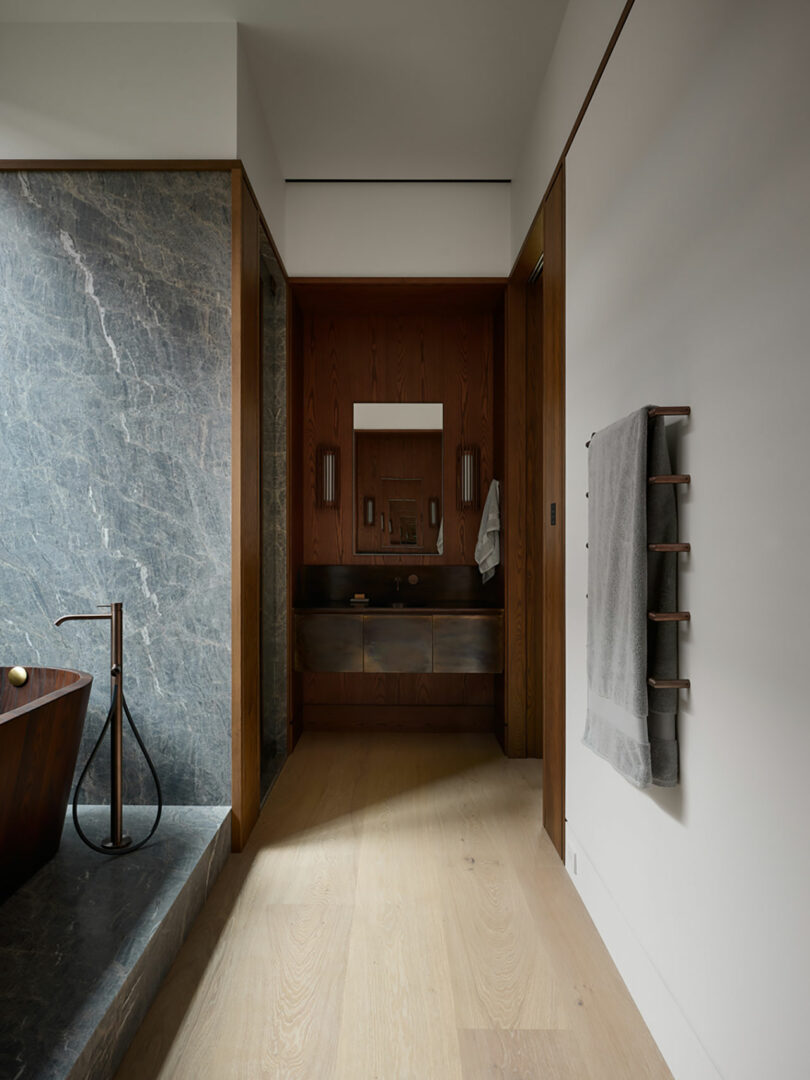 A modern bathroom by Atelier Echelle features a wooden bathtub, stone wall, and elegant wooden vanity. A towel rack is mounted on the right wall, complemented by a large mirror above the sink.