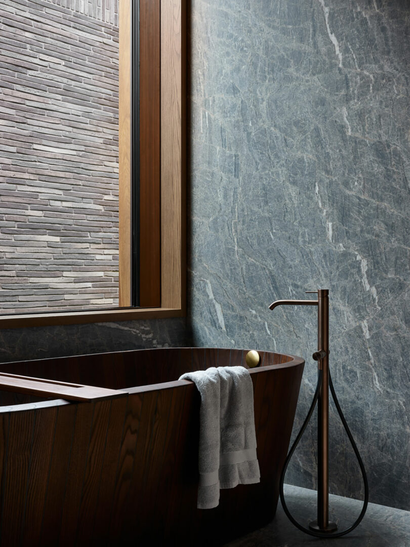 Modern bathroom featuring a dark wooden bathtub, a rolled towel on its edge, and a sleek standalone faucet. A large window with a textured wall view is on the left, enhancing the elegant ambiance.