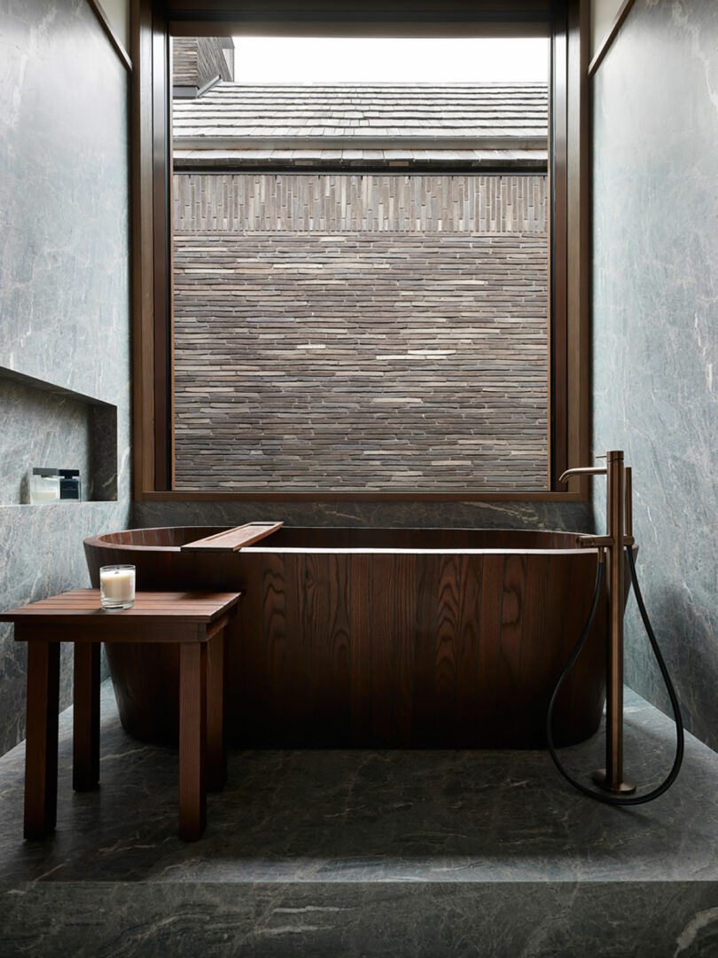 A luxurious bathroom by Atelier Echelle features a dark wood bathtub and a wooden side table with a lit candle, complemented by a floor-mounted faucet. The serene setting is enhanced by a large window overlooking an iconic brick wall, creating an oasis of calm.