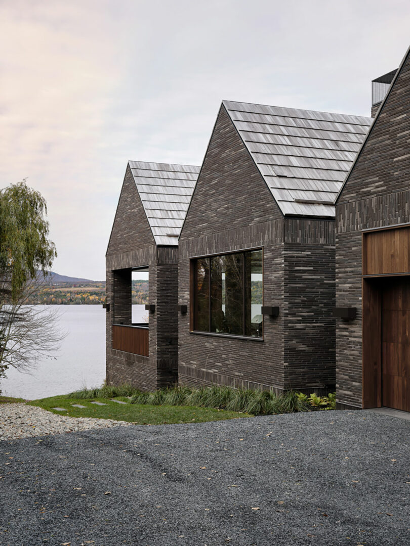 A modern, dark brick house designed by Atelier Echelle features tall, sloped roofs and sits by a lakeside, surrounded by autumn trees and a gravel driveway.