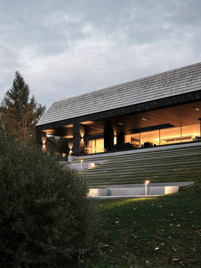 Modern home by Atelier Echelle, featuring a sloped lawn and illuminated steps, with large windows and a wooden roof. The setting is surrounded by trees under a cloudy sky.