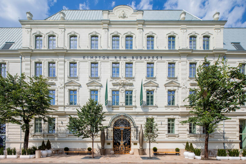 Front view of the Kimpton Budapest Hotel, a large white historic building with ornate architecture, several windows, and small trees in front.