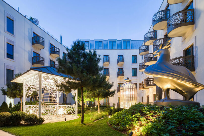 Courtyard of a modern building with rounded balconies, featuring a large metallic bird sculpture and a white, ornate gazebo amidst greenery.