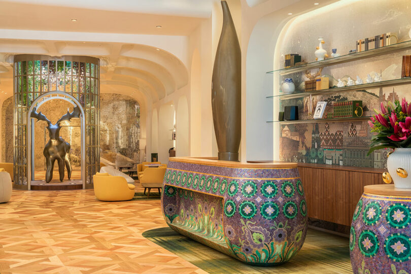 A hotel lobby with colorful decorative tables, a large sculpture, and a deer statue near the entrance. The flooring has a geometric pattern, and there are shelves with various items on display.