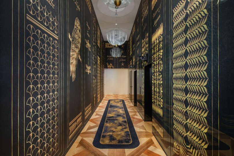 Ornate hallway with dark, patterned walls and gold accents, featuring a long carpet and two glass chandeliers.