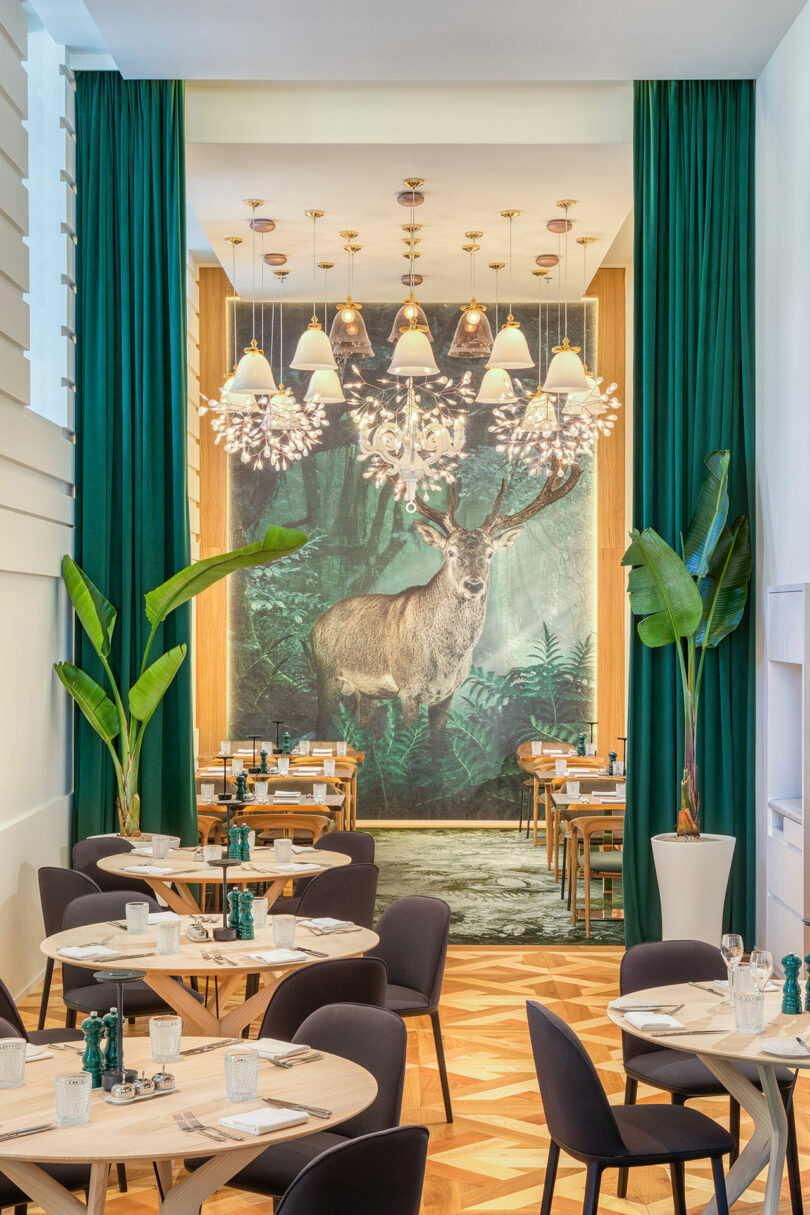 A dining area with wooden tables and chairs, lush plants, and a large deer mural on the back wall, under decorative hanging lights.