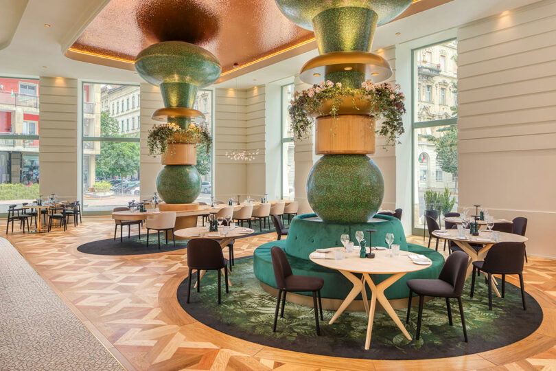 Modern restaurant interior with large green column decorations, wooden tables, and chairs. Natural light filters through large windows, illuminating the geometric patterned floor.