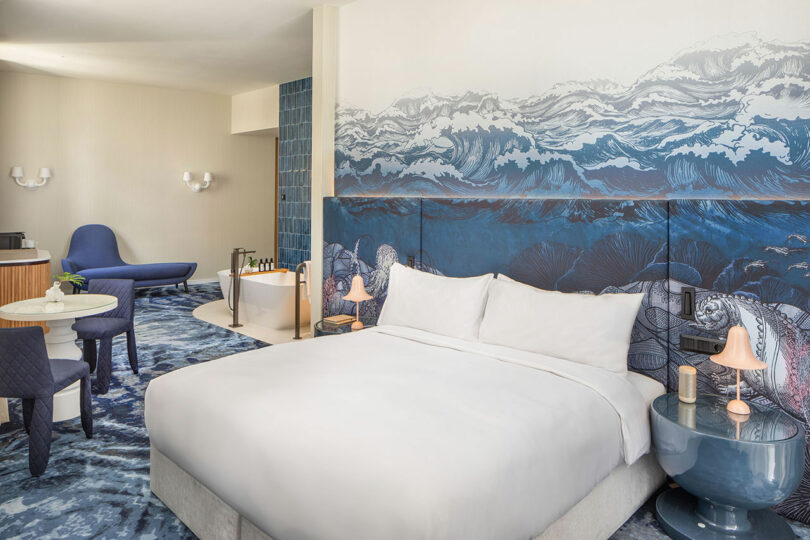 Hotel room with a large bed, ocean-themed decor, and blue accents. Features a seating area, freestanding bathtub, and desk.