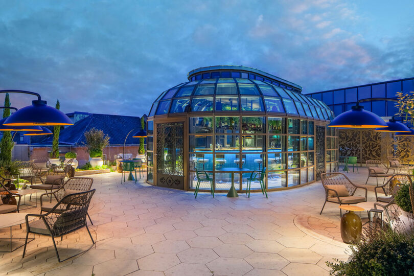 Rooftop terrace with a glass-enclosed bar, outdoor seating, and large hanging lamps, set against a cloudy sky backdrop.