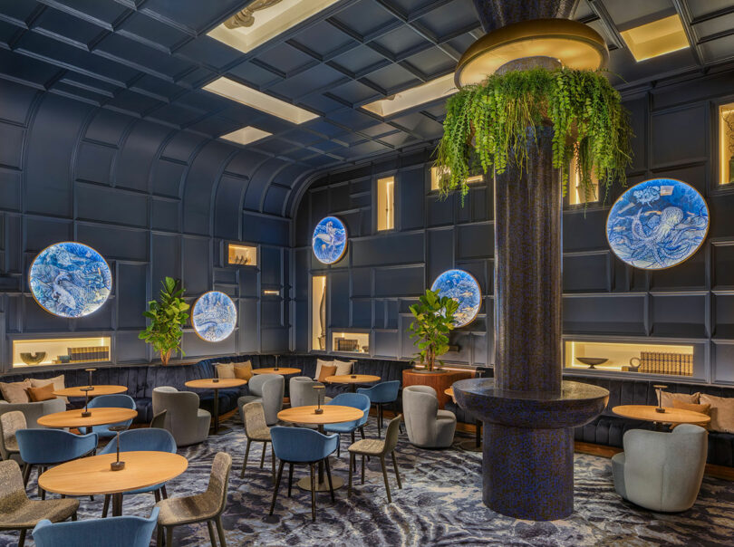 A modern restaurant interior with blue walls, round wooden tables, blue and beige chairs, large decorative plates on the walls, and a central column with greenery.