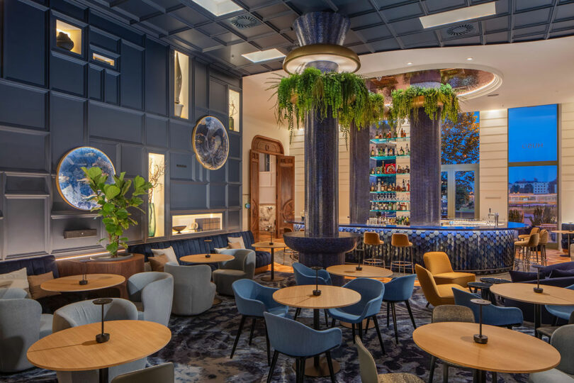 Luxurious bar with modern decor, featuring blue accents, round tables, upholstered chairs, and a central bar counter with hanging plants.