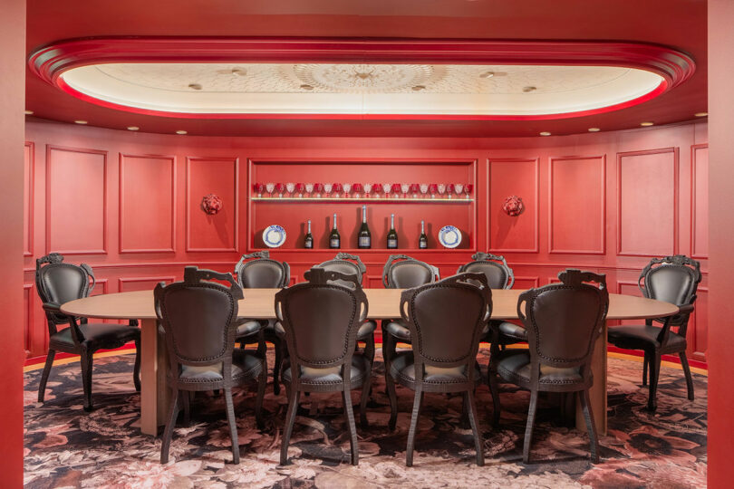 A conference room with a long table, surrounded by black chairs, and a red paneled wall with decorative plates and bottles.