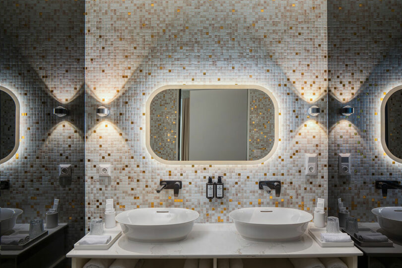 Modern bathroom with two white vessel sinks, a large oval mirror, illuminated by wall sconces. The wall is tiled in a mosaic pattern of light and dark squares.