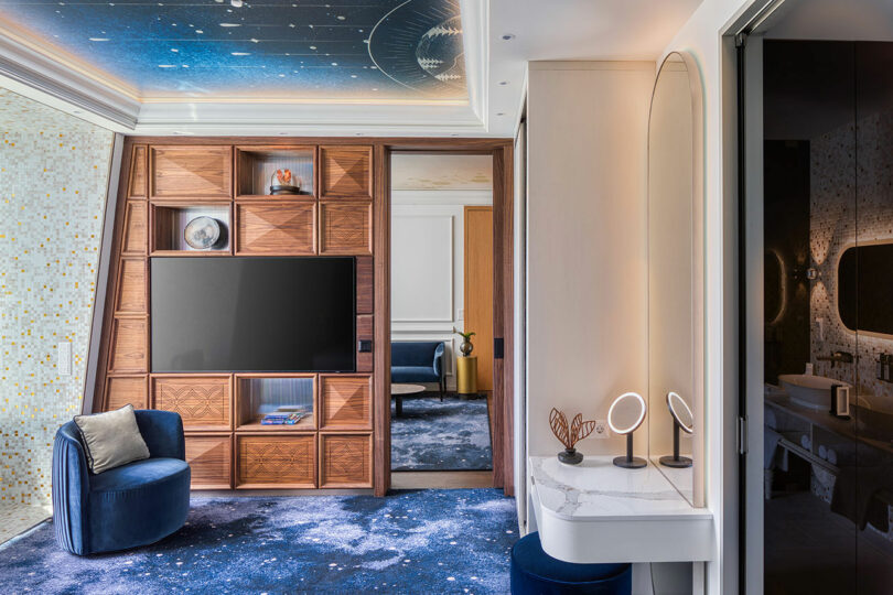 Modern room with a TV on a wooden shelf unit, a blue velvet chair, a dressing table with mirrors, and a starry ceiling design. Doorway leads to an adjacent room.