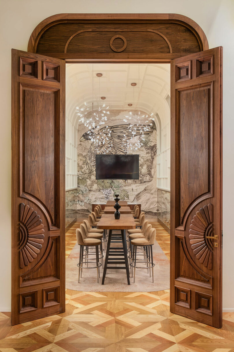 Open wooden doors lead to a conference room with a long table, beige chairs, a large screen, and a decorative ceiling light fixture. The room features intricate wall artwork.