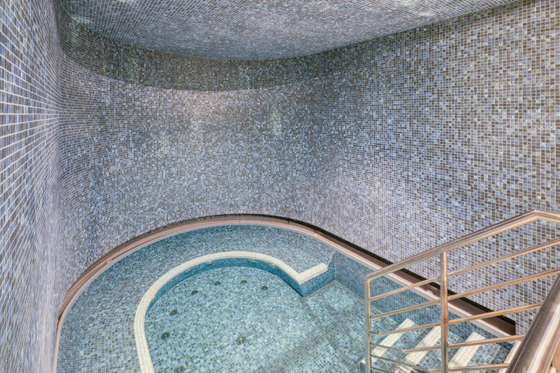 A mosaic-tiled indoor spa area with a circular hot tub is surrounded by curved walls. Stairs with metal railings lead into the spa.