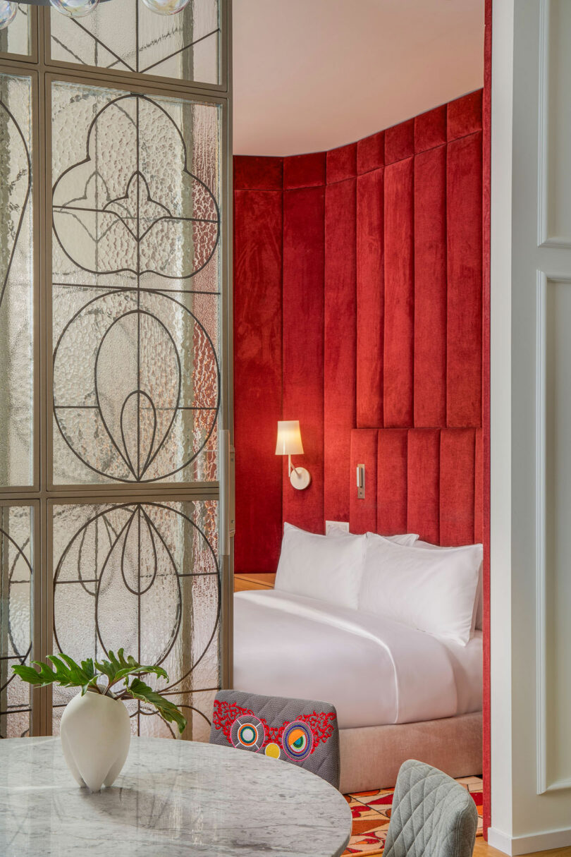 Luxurious hotel room with a plush red velvet headboard, white bedding, decorative glass partition, and a round marble table with a white vase.