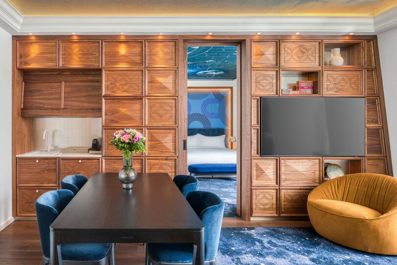 Modern hotel suite with a dining table and chairs, a vase with flowers, wooden shelving, a wall-mounted TV, and an orange chair. Bedroom visible through a sliding door.