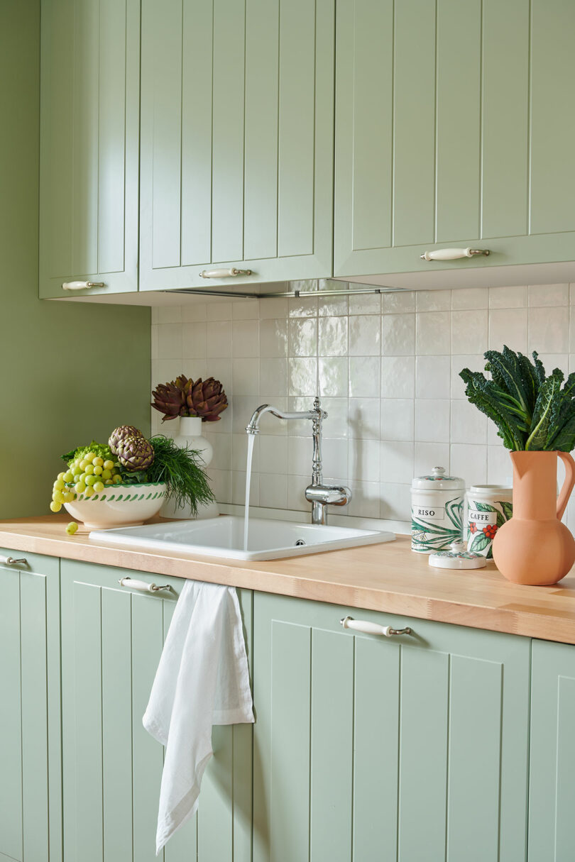 Una cocina con gabinetes de color verde claro, fregadero blanco y encimera de madera. En el mostrador hay productos frescos, como uvas, piña y col rizada. Una toalla blanca cuelga junto al lavabo.