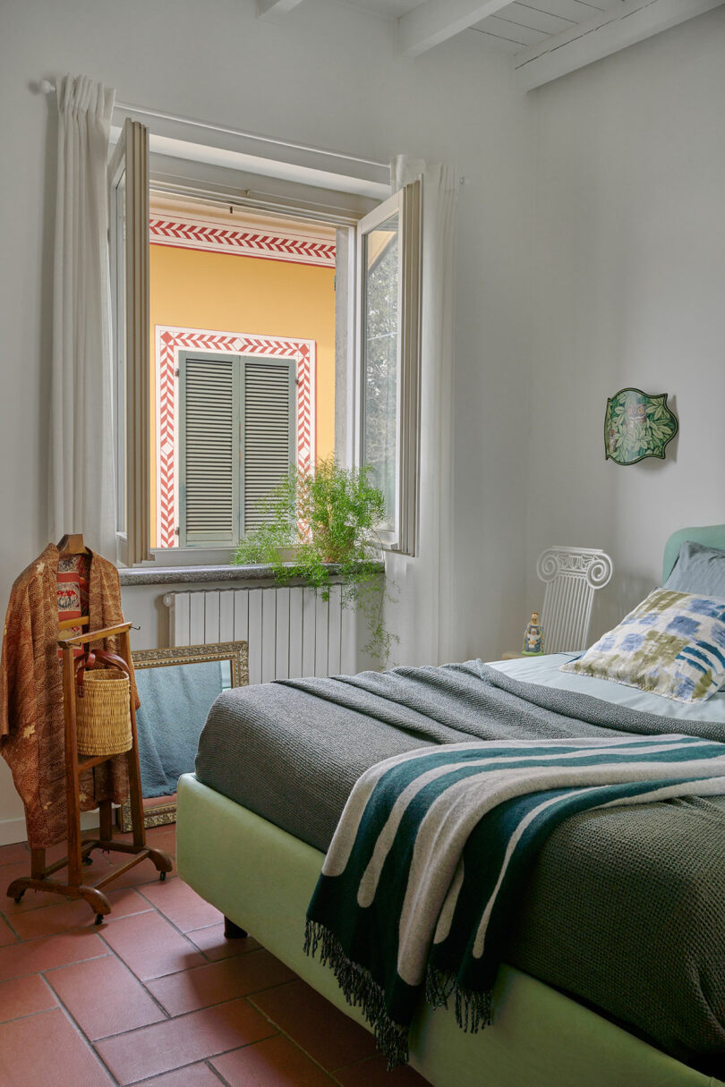 Un dormitorio con una ventana abierta, paredes de colores claros, una cama verde con almohadas y mantas estampadas, una maceta en el alféizar y una silla y percheros con ropa cerca de la cama.