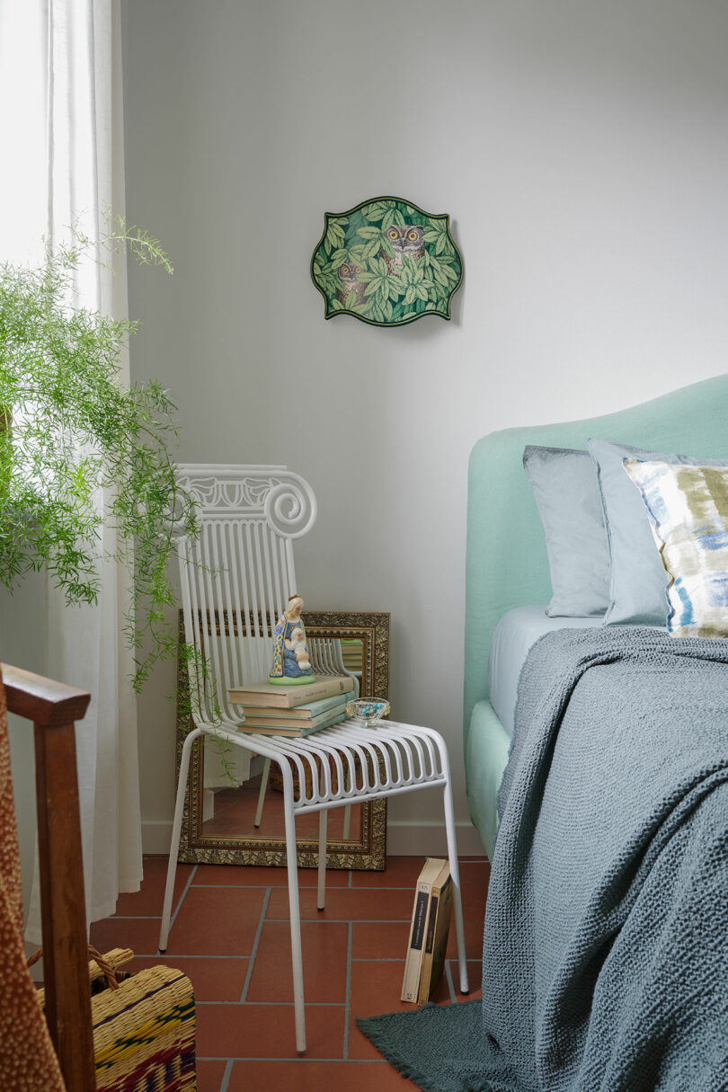 Un dormitorio acogedor con una silla blanca con libros y decoración, junto a una cama verde menta con ropa de cama de color gris azulado. Una frondosa obra de arte en la pared cuelga arriba y una planta en maceta está cerca.