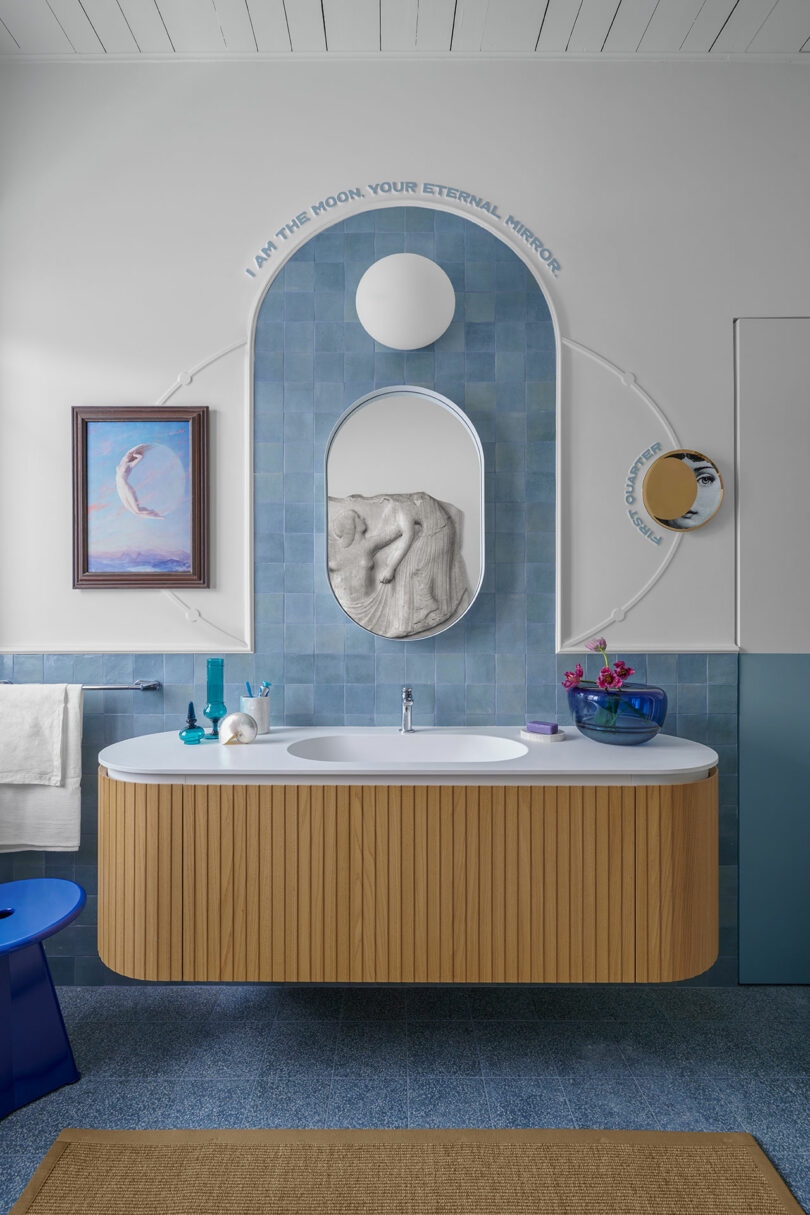 Cuarto de baño moderno con tocador curvo de madera, azulejos azules y espejo redondo. La inscripción en la pared dice: "Soy la luna, tu espejo eterno." La decoración incluye obras de arte y un espejo pequeño.