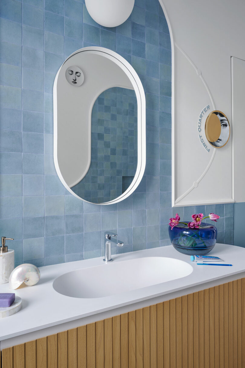 Baño con tocador de madera, espejo ovalado, azulejos azules y encimera blanca. En la encimera, junto al jabón y una concha, hay un pequeño cuenco con flores rosas.