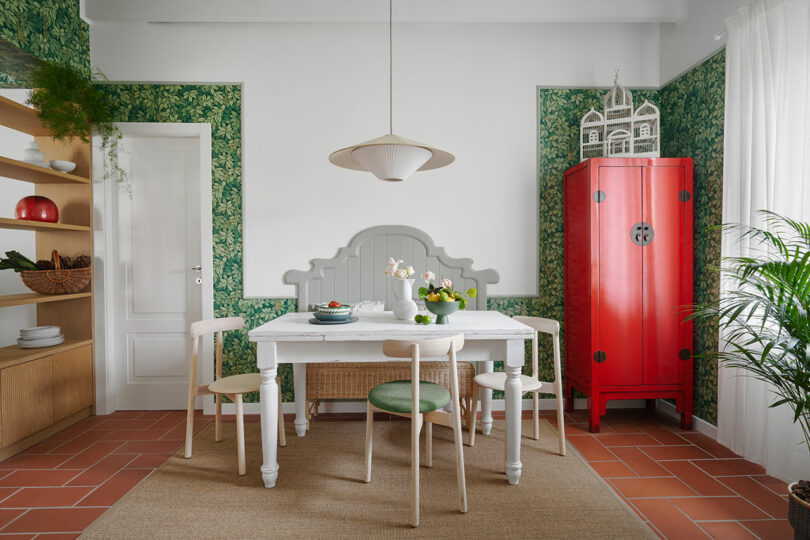 Un comedor con una mesa blanca, sillas verdes, un mueble rojo y papel pintado con motivos verdes. Una lámpara colgante cuelga encima de la mesa y una alfombra tejida sobre el suelo de baldosas de ladrillo.