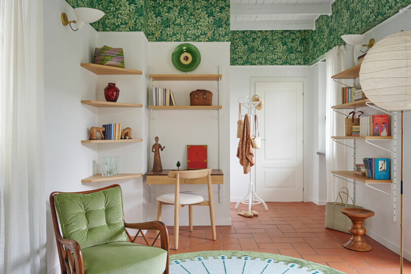 Una acogedora habitación con papel tapiz de follaje verde, un pequeño escritorio con una silla, estantes abiertos con libros y decoración, un piso de madera y una silla con cojines verdes en primer plano.