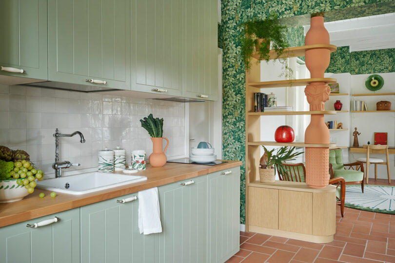 Una cocina con gabinetes verdes, encimera de madera, fregadero blanco y protector contra salpicaduras de azulejos. Un estante con elementos decorativos se encuentra junto a una pared empapelada con un patrón de hojas, lo que da paso a una acogedora habitación.