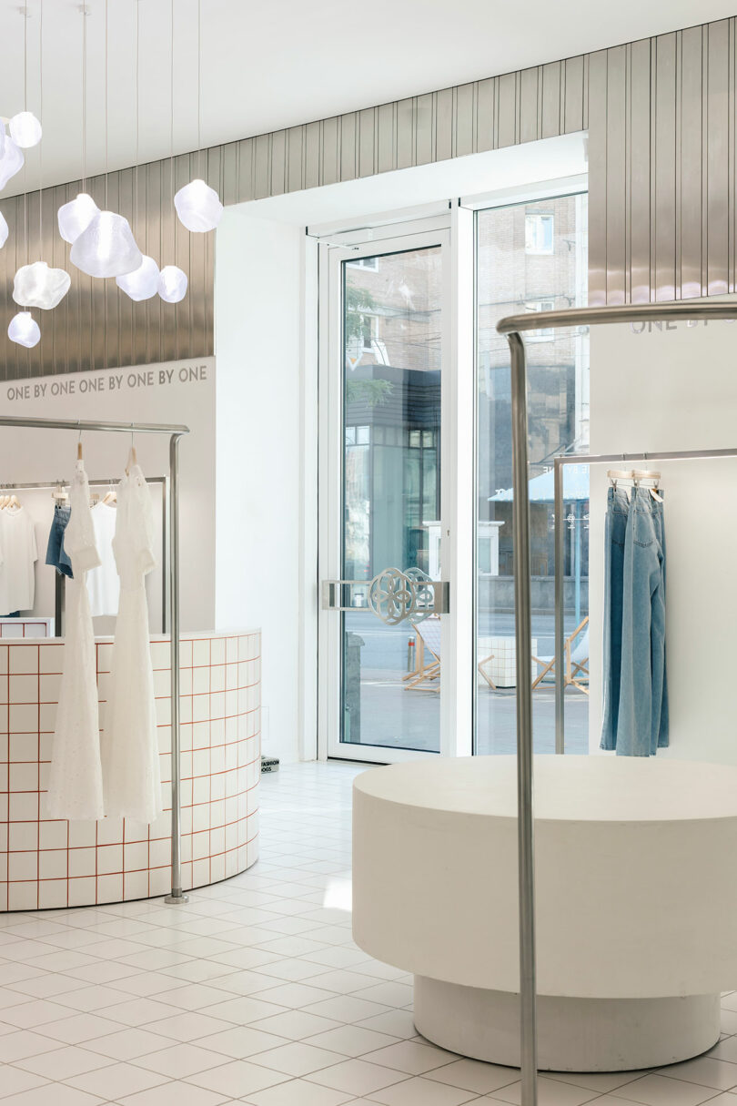 The minimalist clothing store, One by One, features white walls and floors. Hanging garments include dresses and jeans, while modern lighting fixtures illuminate the space. A round display table stands centrally, enhancing the sleek aesthetic.