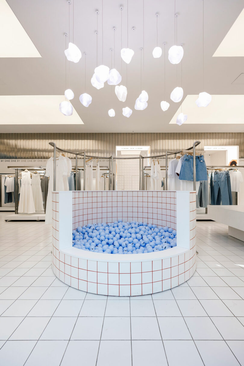 The modern clothing store interior, with its white and blue theme, invites customers to explore, One by One. A central display features a circular pit filled with blue balls, while clothes hang neatly around the perimeter under elegant white light fixtures.