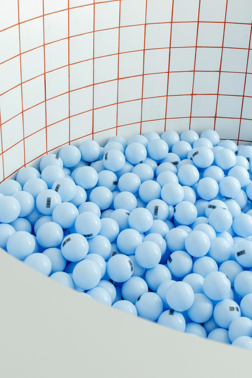 A pool filled with numerous light blue balls, each marked with a barcode, lies against a background of white tiles with red grid lines.