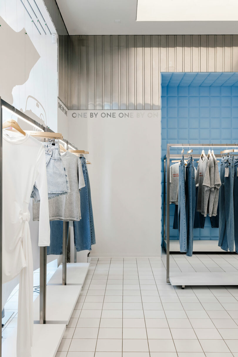 A modern clothing store interior with white tile flooring showcases a variety of denim apparel and white tops on racks, displayed one by one against a blue and white backdrop.