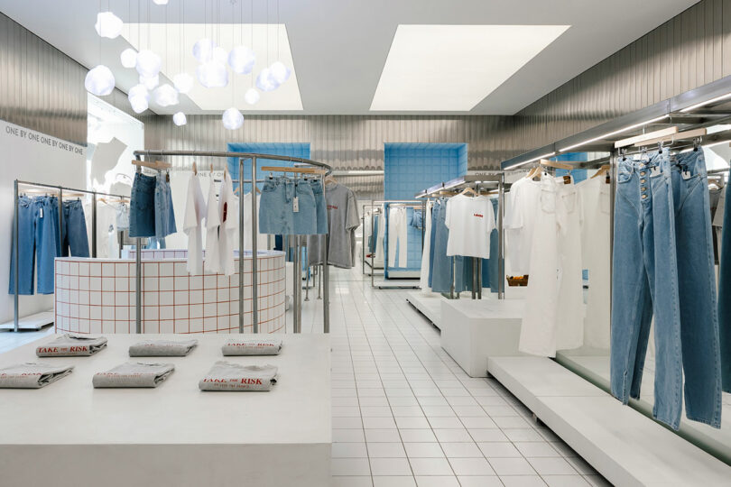 A modern clothing store interior designed with a minimalist touch features white walls, checkered displays, and racks filled with denim jeans and white tops. Bright overhead lights illuminate the space, inviting customers to discover each stylish piece One by One.