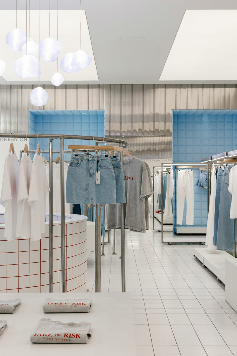 The One by One clothing store interior boasts a chic white and blue decor. Stylish displays feature white t-shirts, light blue denim skirts, and folded gray shirts on sleek white grids. Modern lighting and reflective surfaces complete the look for a contemporary shopping experience.