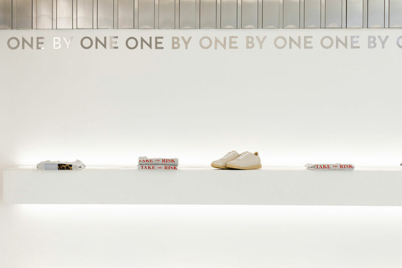 A minimalist store display features white sneakers, folded shirts, and books on a white shelf against a pristine white wall adorned with a subtle "One by One" text pattern, creating an elegantly unified aesthetic.