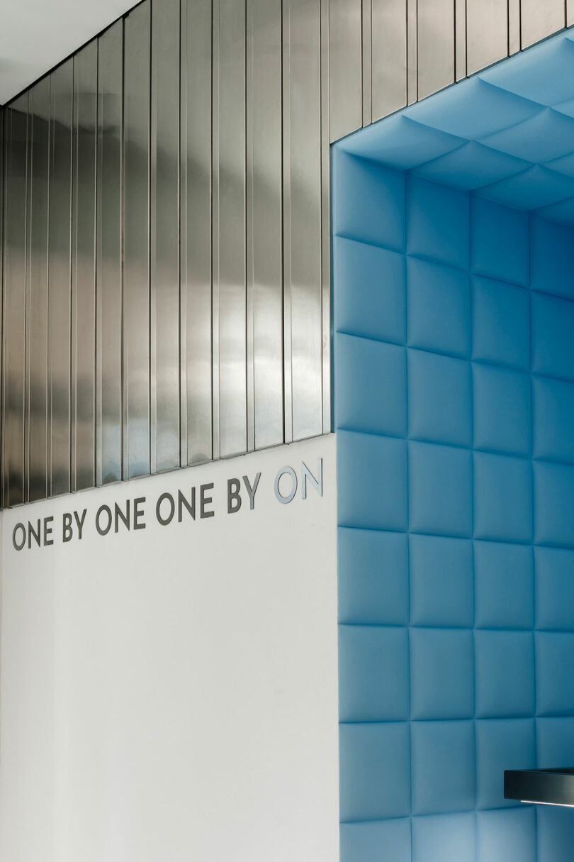 Modern interior with silver metallic walls and a blue cushioned alcove. The text "ONE BY ONE, ONE BY ONE" seamlessly integrates into the design, echoing throughout the space.