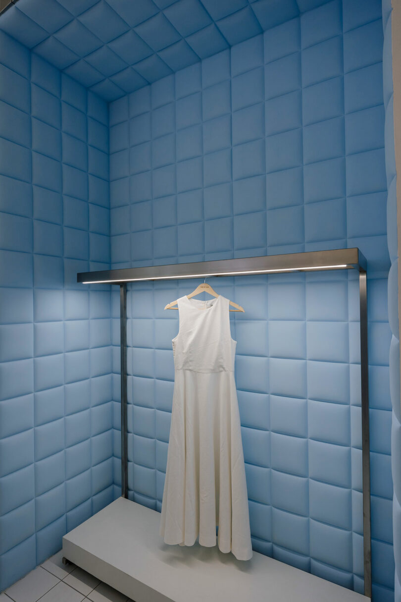 A white, sleeveless dress from One by One hangs gracefully on a wooden hanger inside a blue padded dressing room with soft lighting.