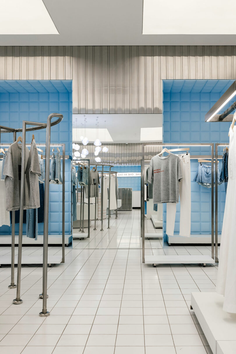 The One by One clothing store features blue padded walls and sleek metal racks displaying casual wear, set against pristine white tiled flooring.