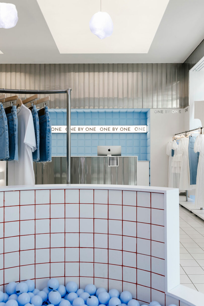 The clothing store interior, exuding minimalist elegance, showcases racks of jeans and a sleek white counter. A playful ball pit with blue balls complements the light blue tiled wall adorned with a sign reading "One By One," creating a harmonious shopping atmosphere.