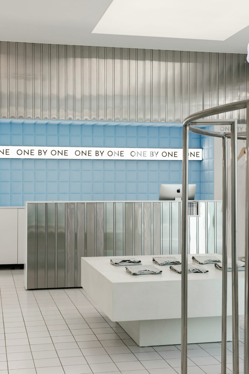 Modern retail store interior with metallic walls and a blue grid panel. The sleek white counter displays clothing alongside a computer. In the background, the store name "One by One" is elegantly showcased, adding to the chic ambiance.