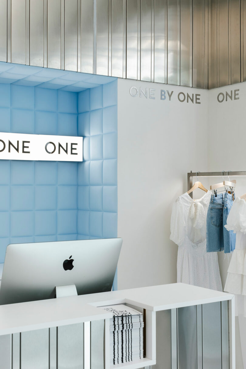 A retail store interior with a light blue wall adorned with the text "One by One," showcasing clothing on display. A computer rests on a sleek white counter, adding to the modern ambiance.