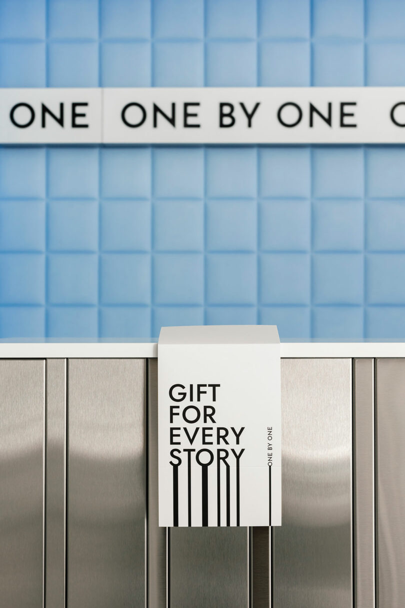 A sign with "Gift for Every Story" hangs over a metal surface, perfectly complementing the tiled blue wall and offering a nod to the phrase "One by One" in the background.