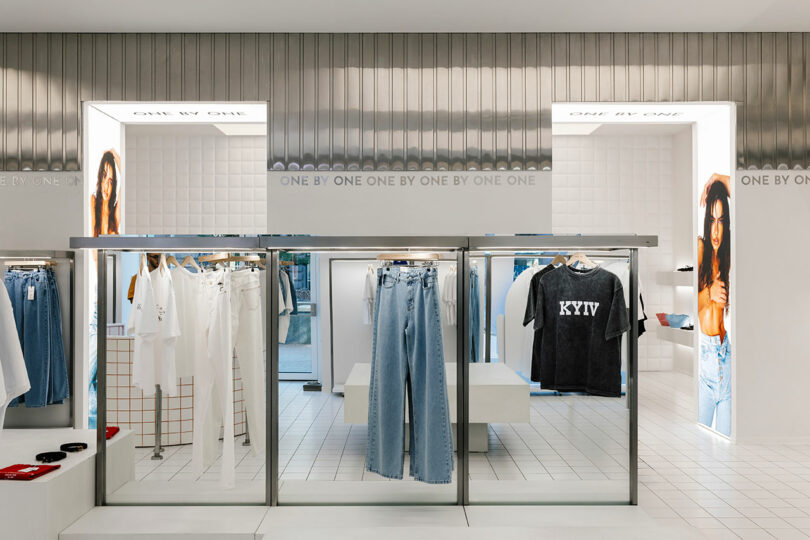 The clothing store interior features a sleek white tile floor and metallic accents. Racks display shirts and jeans, arranged one by one for easy browsing. Large photos of models hang on the walls, adding a touch of inspiration to your shopping experience.