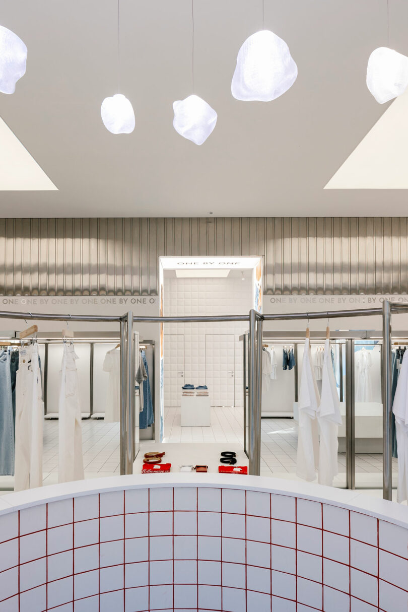 A modern clothing store interior with a minimalist design, where style is curated one by one. White walls and a red grid counter are in the foreground, while racks of neatly hung clothes are visible in the background.