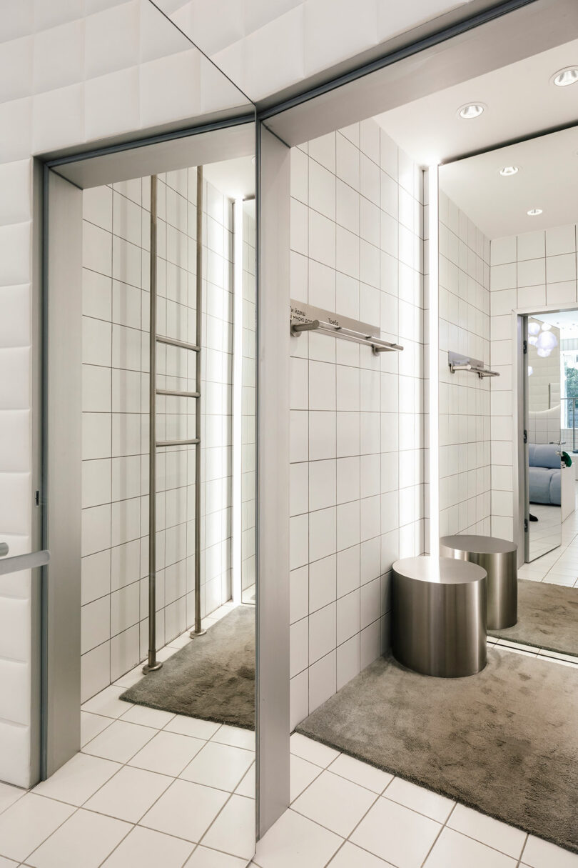 Modern dressing room with white tiled walls, circular stools, floor-to-ceiling mirrors, and metal rails. Step in One by One to experience the elegance of this meticulously crafted space.