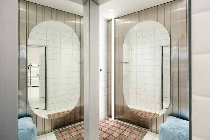 A modern changing room boasts two large oval mirrors on tiled walls, illuminated by vertical lights one by one, and featuring gray and red plaid rugs near blue cushioned stools.