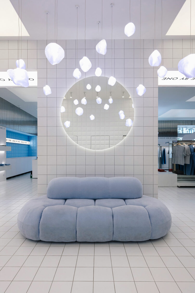 The modern showroom features a soft blue sofa, a large circular mirror, and white spherical pendant lights aligned one by one against the tiled wall and floor.